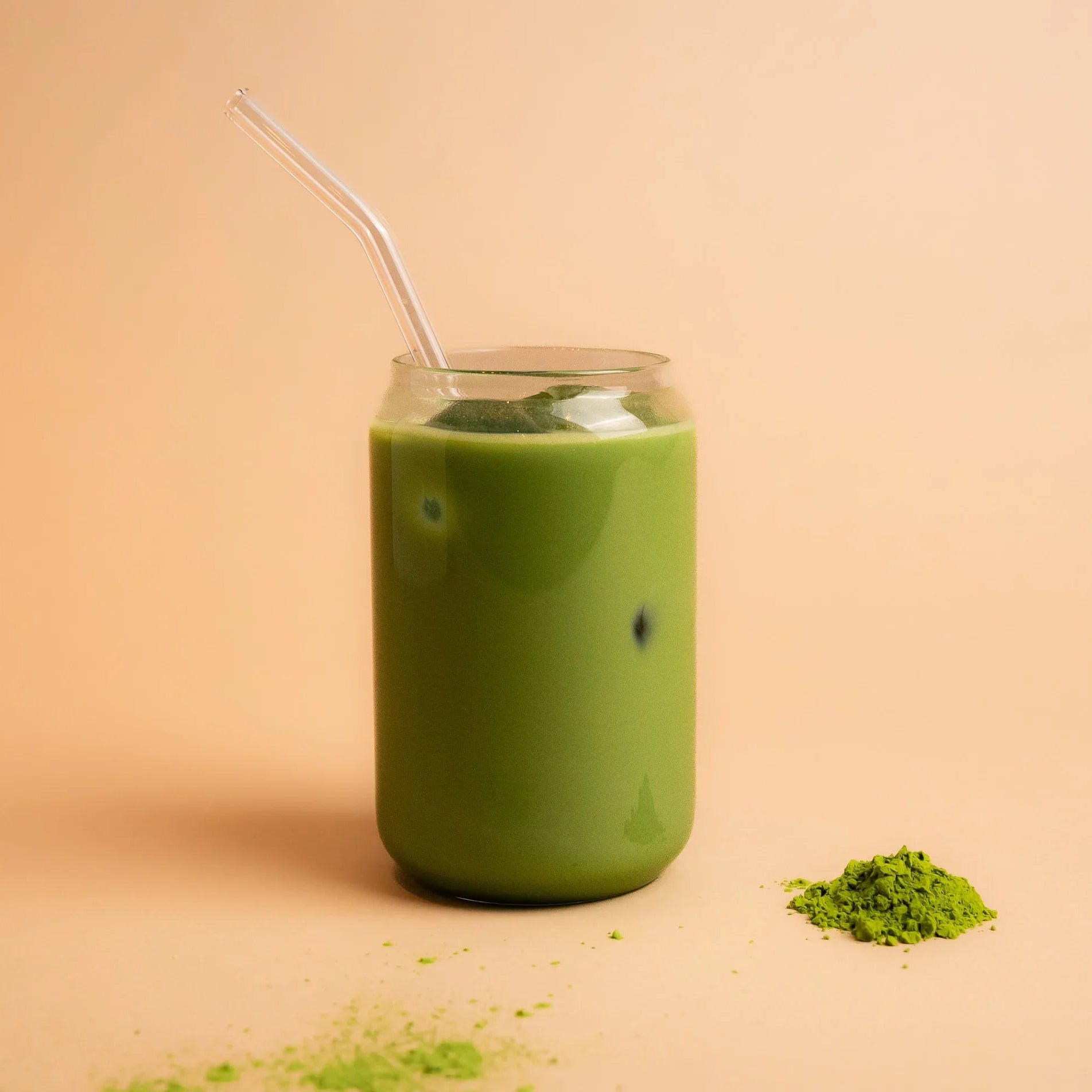 Iced matcha latte in a glass with a straw beside matcha powder on a neutral background